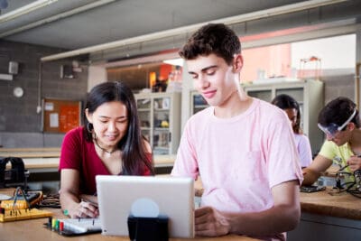 Two students work together at a desk with a laptop in a classroom, while two others work in the background.