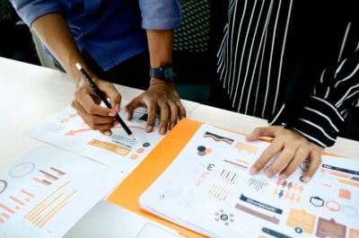 Two people reviewing documents with graphs and charts at a table. One person holds a pencil, and both have their hands on the papers.