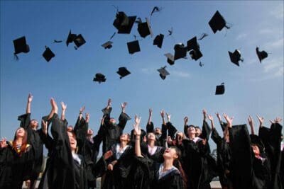 Graduates in caps and gowns celebrate by tossing their caps into the air against a clear blue sky.