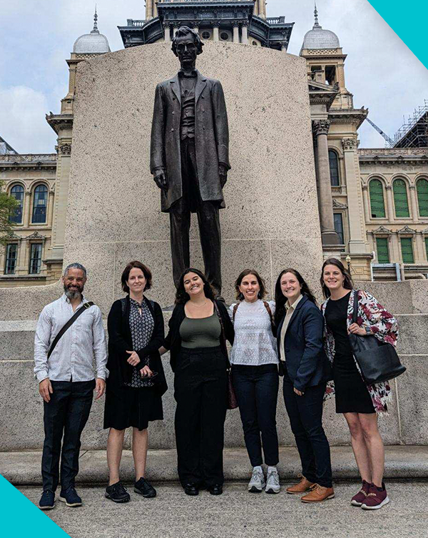 Six people standing in front of a large statue outside a historic building.