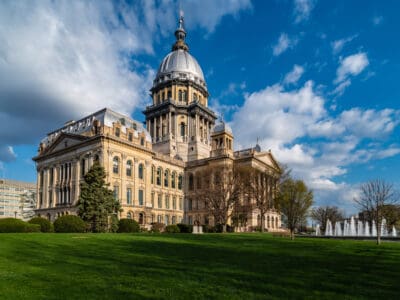 The Illinois State Capitol building with its distinctive dome, surrounded by a well-maintained lawn and trees under a partly cloudy sky.