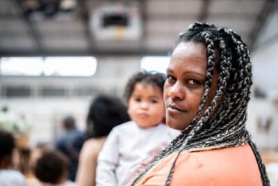 A woman with braided hair holds a young child in her arms in an indoor setting. Other people are blurred in the background.