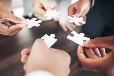 Five people each holding a white puzzle piece, gathering in a circle over a wooden table surface.