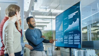 Four people in a modern office discuss data on a large screen, showing graphs and analytics.