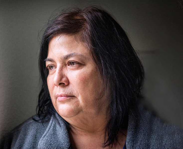 A woman with dark shoulder-length hair looks out a window, wearing a grey top.