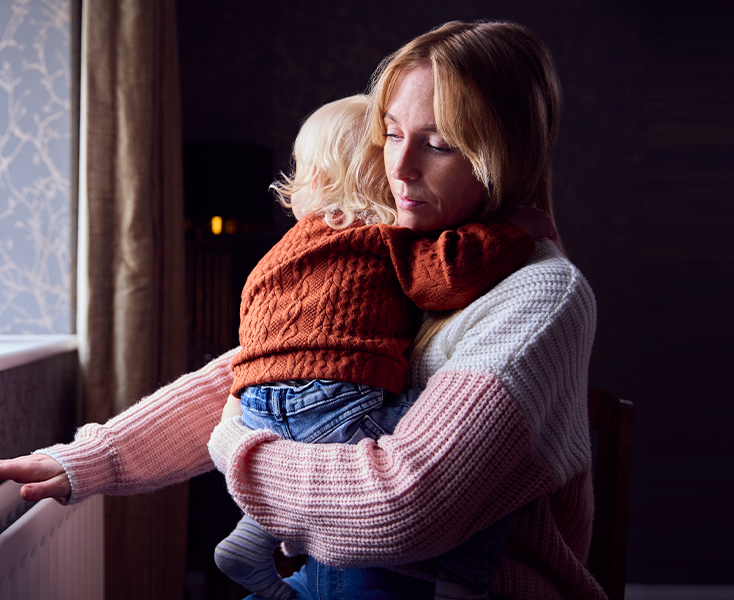 A woman in a knitted sweater holds a child with blonde hair, wearing a rust-colored sweater, in a dimly lit room.