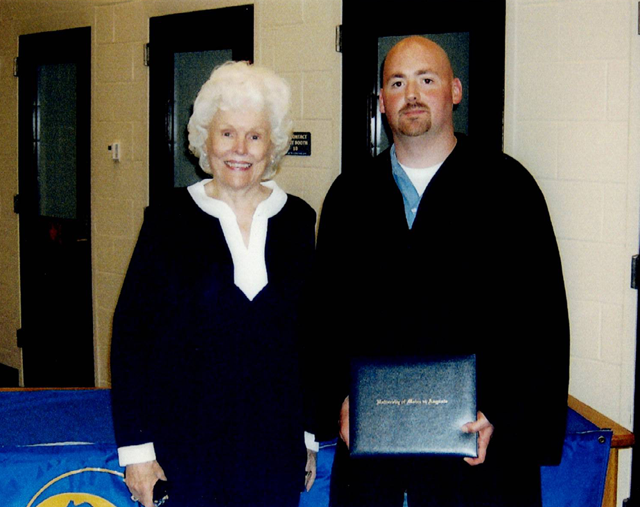 A man in a graduation gown stands next to an elderly woman in a hallway. The man holds a diploma.