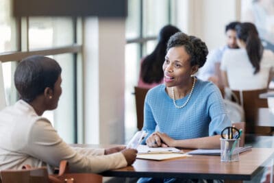 Two women sit at a table in an office setting, engaged in conversation. One woman has a notepad. The atmosphere is professional and friendly.