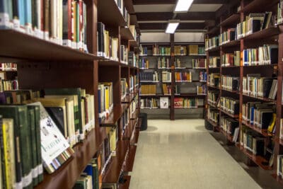 A library aisle with wooden shelves filled with various books, well-lit by overhead fluorescent lights.