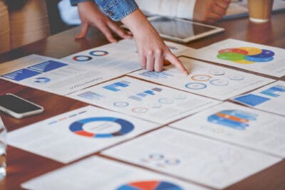A person points to a chart on a table covered with various business graphs, charts, and documents.