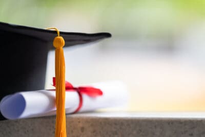 Graduation cap with yellow tassel and a rolled diploma tied with a red ribbon on a stone surface, with a blurred green background.