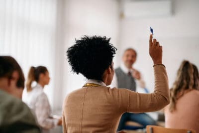 Student with curly hair raises hand in a classroom while holding a pen.