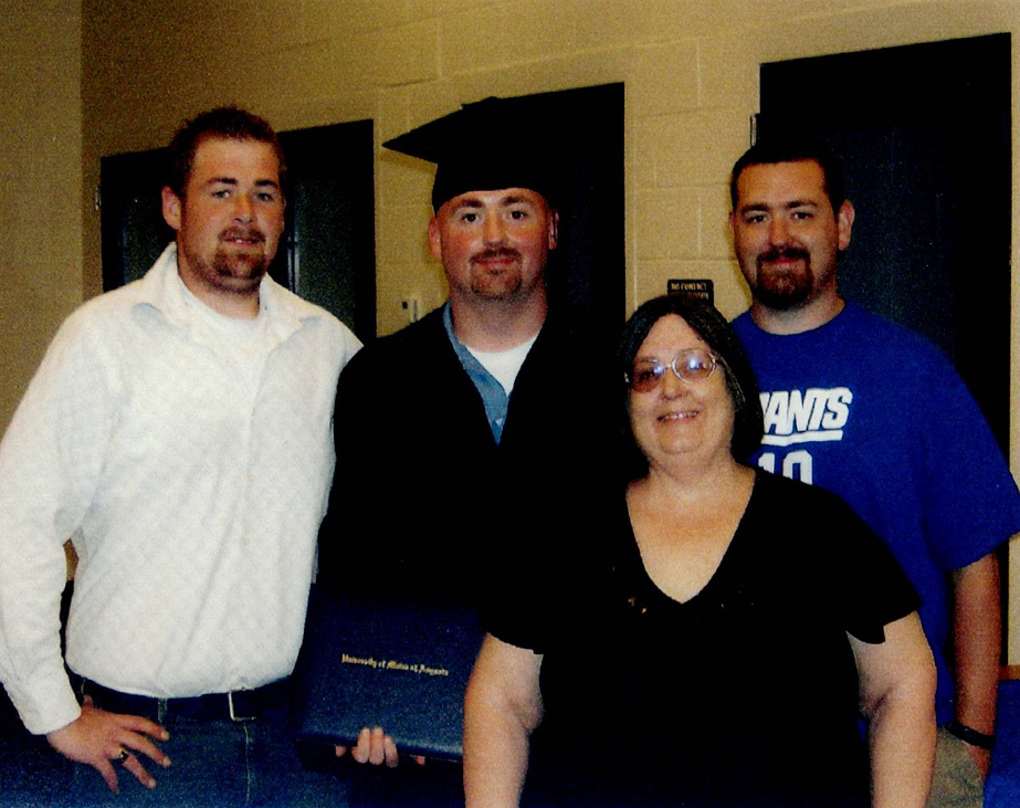 A graduate in a cap and gown holds a diploma, posing with three people indoors.
