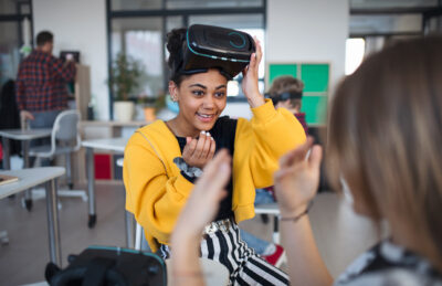 Young woman in a yellow sweater, sitting and wearing a VR headset, interacting with another person in a classroom setting.