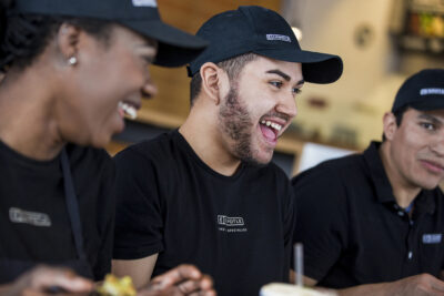 Three employees wearing black shirts and caps with a logo, sitting and smiling together indoors.