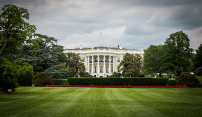 A distant view of the White House with a well-manicured lawn and surrounding trees under a cloudy sky.