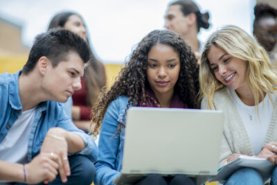 Three young adults sit outdoors, focused on a laptop screen. The person in the middle uses the laptop, while the others look on. More people are in the background.