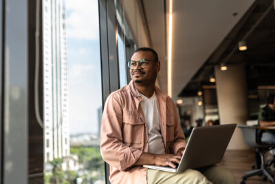 A person sits by a large window in an office setting, working on a laptop and gazing outside thoughtfully.