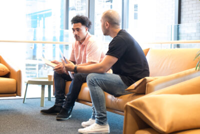 Two men sitting on a brown sofa, engaged in conversation, one holding a tablet. They are in a brightly lit, modern indoor space with large windows in the background.