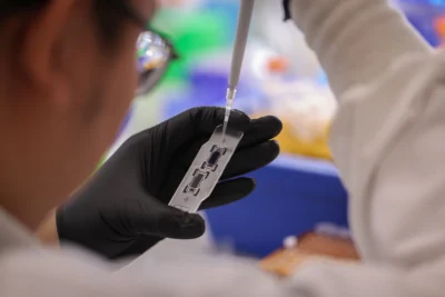 A person wearing black gloves uses a pipette to add liquid to a small transparent device in a laboratory setting.
