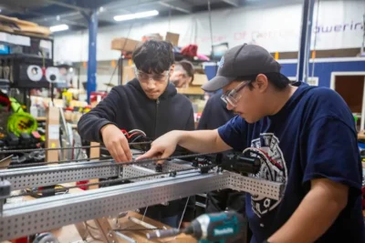 Two students wearing safety goggles assemble a metal robotic structure in a workshop filled with tools and equipment.