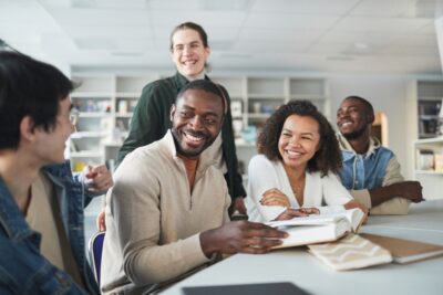 Five people sit and stand around a table in a bright room, smiling and laughing, with open books and notebooks in front of them.
