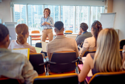 A woman stands in front of a seated group in a classroom setting, engaging with them. She is smiling and holding a marker, while participants listen and take notes.
