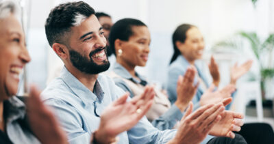 A group of people sitting and clapping, smiling and appearing engaged, in what looks like a professional or educational setting.