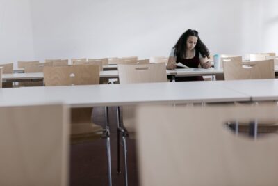 A person with curly hair reads at a desk in an empty classroom with rows of wooden chairs and white tables.