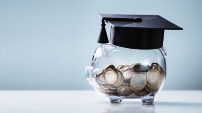 A clear piggy bank filled with coins wearing a graduation cap, symbolizing saving for education.