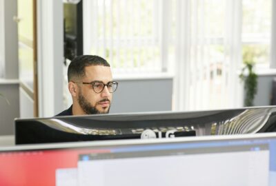 Man with glasses working at a computer in a bright office environment. Two monitors are visible.