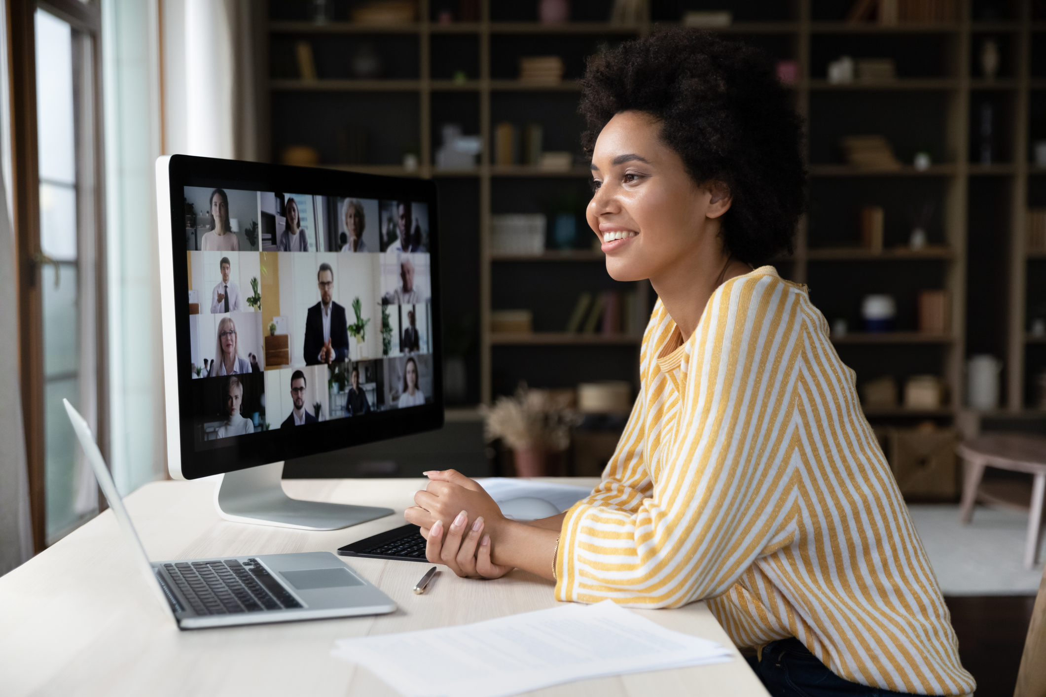 A woman sits at a desk, smiling at a computer screen displaying a video conference with multiple participants. Papers, a pen, and a laptop are on the desk.