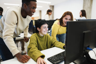 Three students gather around a computer monitor in a classroom, smiling and looking at the screen. Other students are visible in the background.