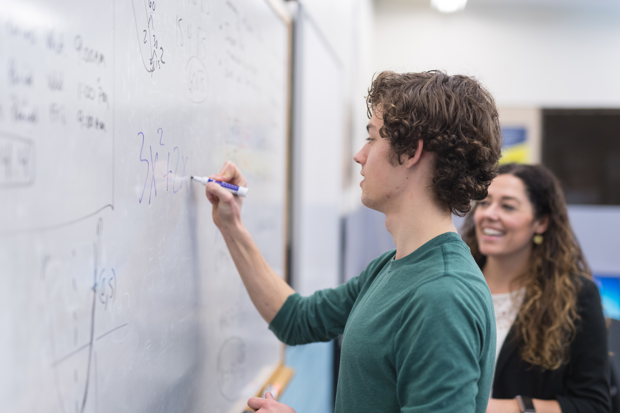A young man writes a math equation on a whiteboard while a woman stands nearby smiling.