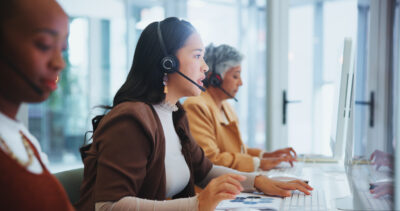 Three women sit at desks wearing headsets, working on desktop computers in a modern office environment.