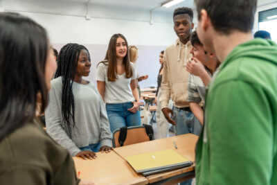 A group of students stand and talk around a desk in a classroom, with notebooks and folders on the table.
