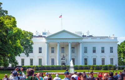 The White House, a white neoclassical building with an American flag on top, viewed from outside the front fence with a crowd of people in the foreground.