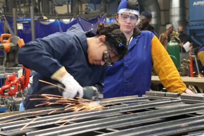 Two people in safety gear work in a metal workshop; one uses a grinder, producing sparks, while the other observes closely.