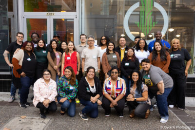 A diverse group of people pose together for a group photo on a sidewalk in front of a storefront with glass doors and windows.