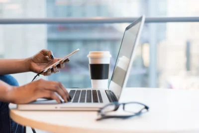 A person types on a laptop with one hand while holding a smartphone in the other; a coffee cup and eyeglasses rest on the table nearby.