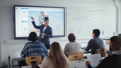 An instructor in business attire presents data on a large screen to a group of students seated in a classroom with laptops, while diagrams are visible on a whiteboard.