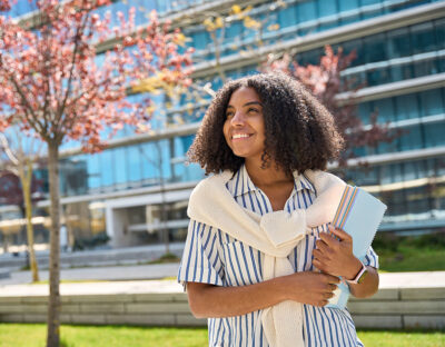 A young person stands outdoors in front of a modern glass building, holding books and smiling, with blooming trees in the background.