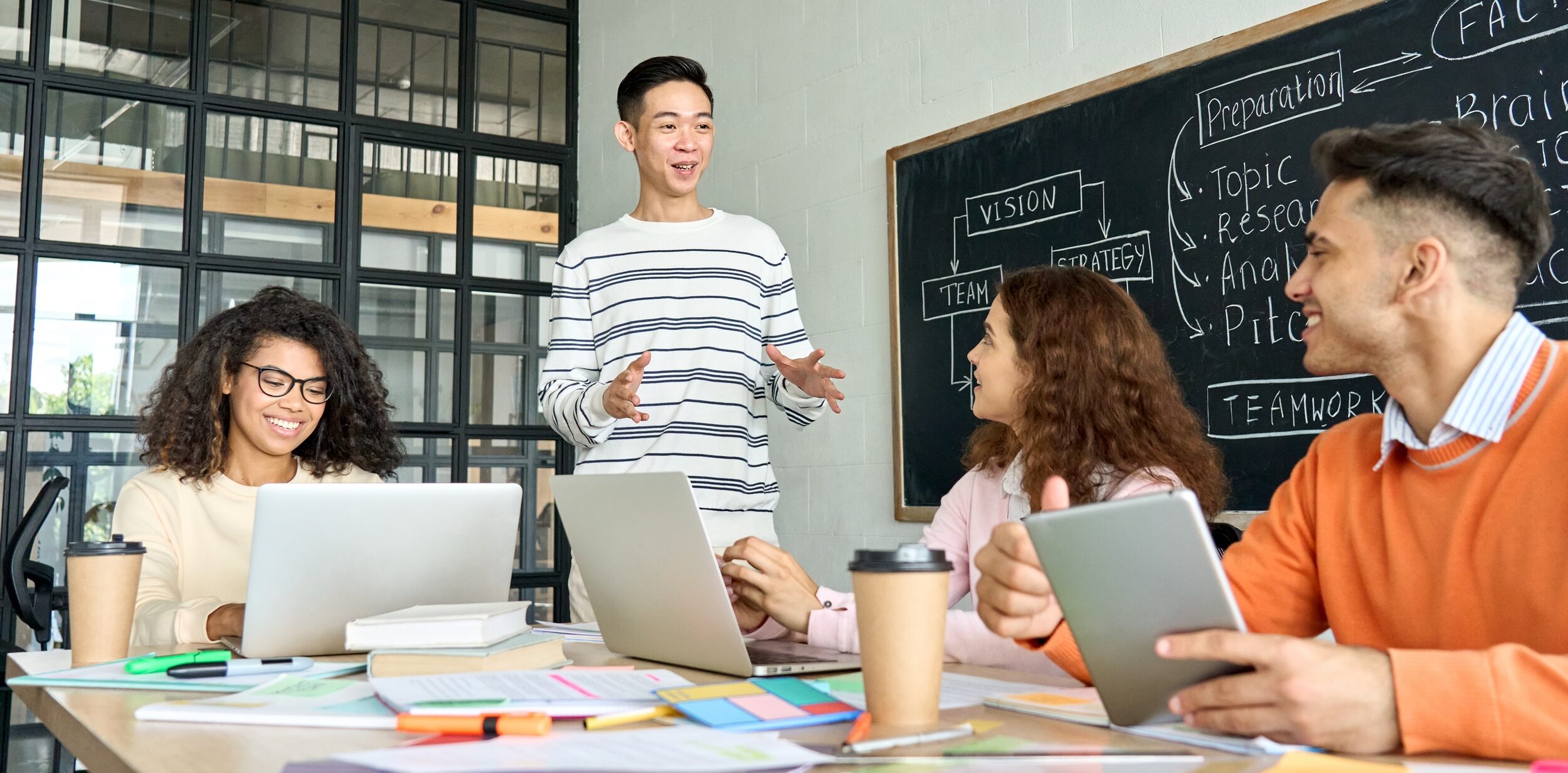 Four people have a discussion in a modern office, with laptops, tablets, and papers on the table. A man stands speaking, while three others sit and listen. A chalkboard with notes is behind them.