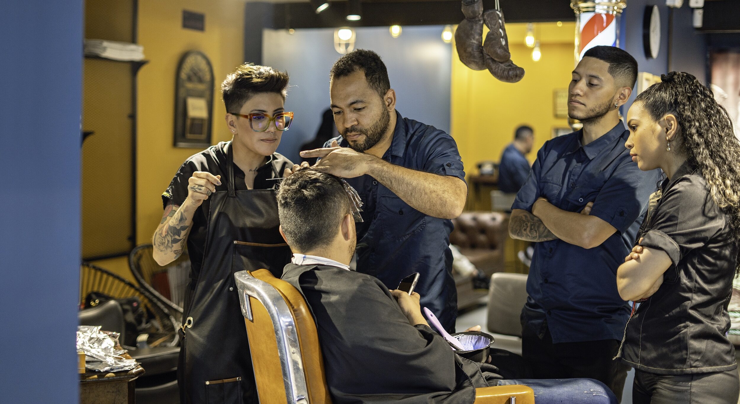 Barbers and stylists work together cutting a client's hair in a modern barbershop, while two other staff members observe closely.