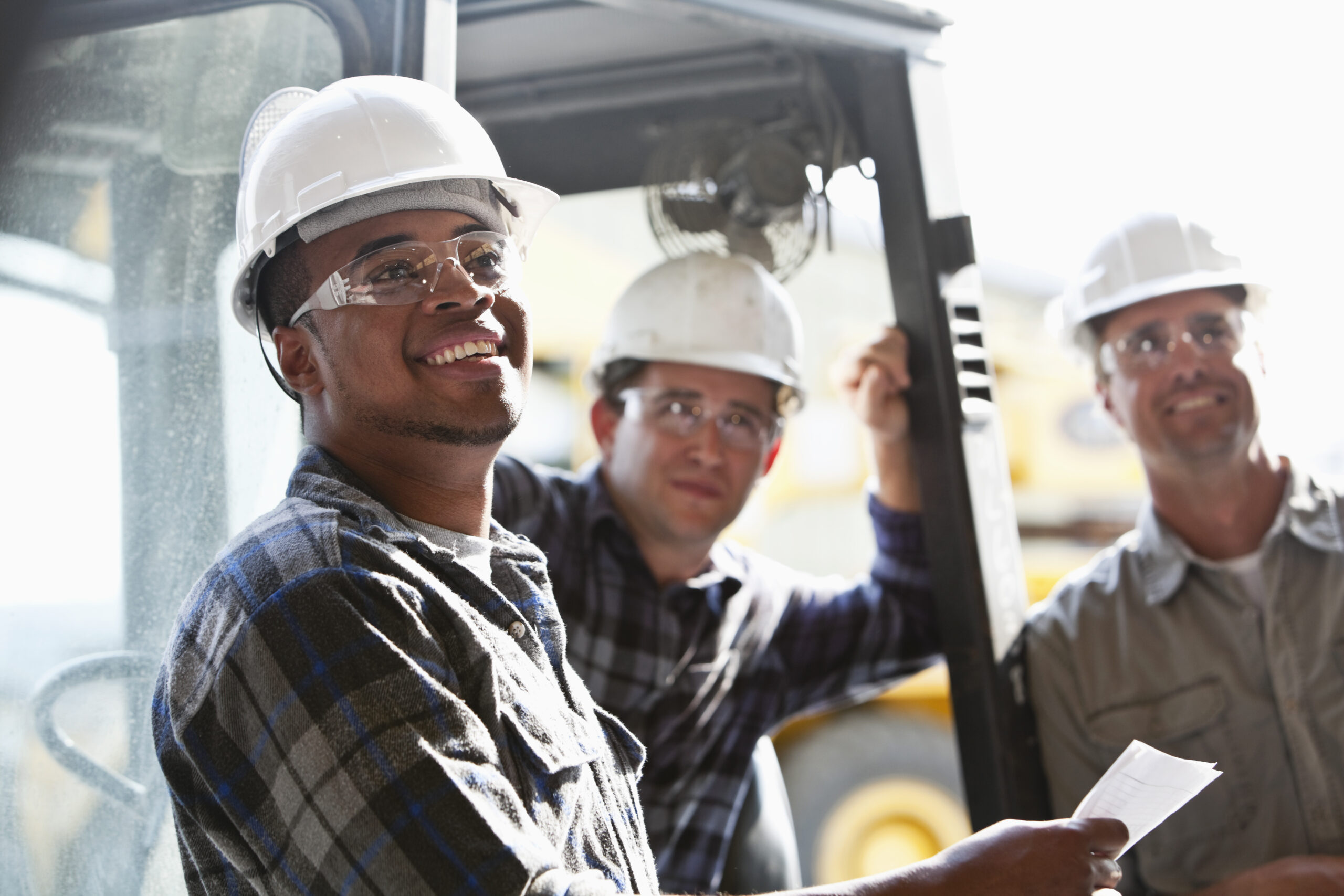 Three construction workers wearing hard hats and safety glasses stand together outdoors, one holding a piece of paper, with construction equipment visible in the background.