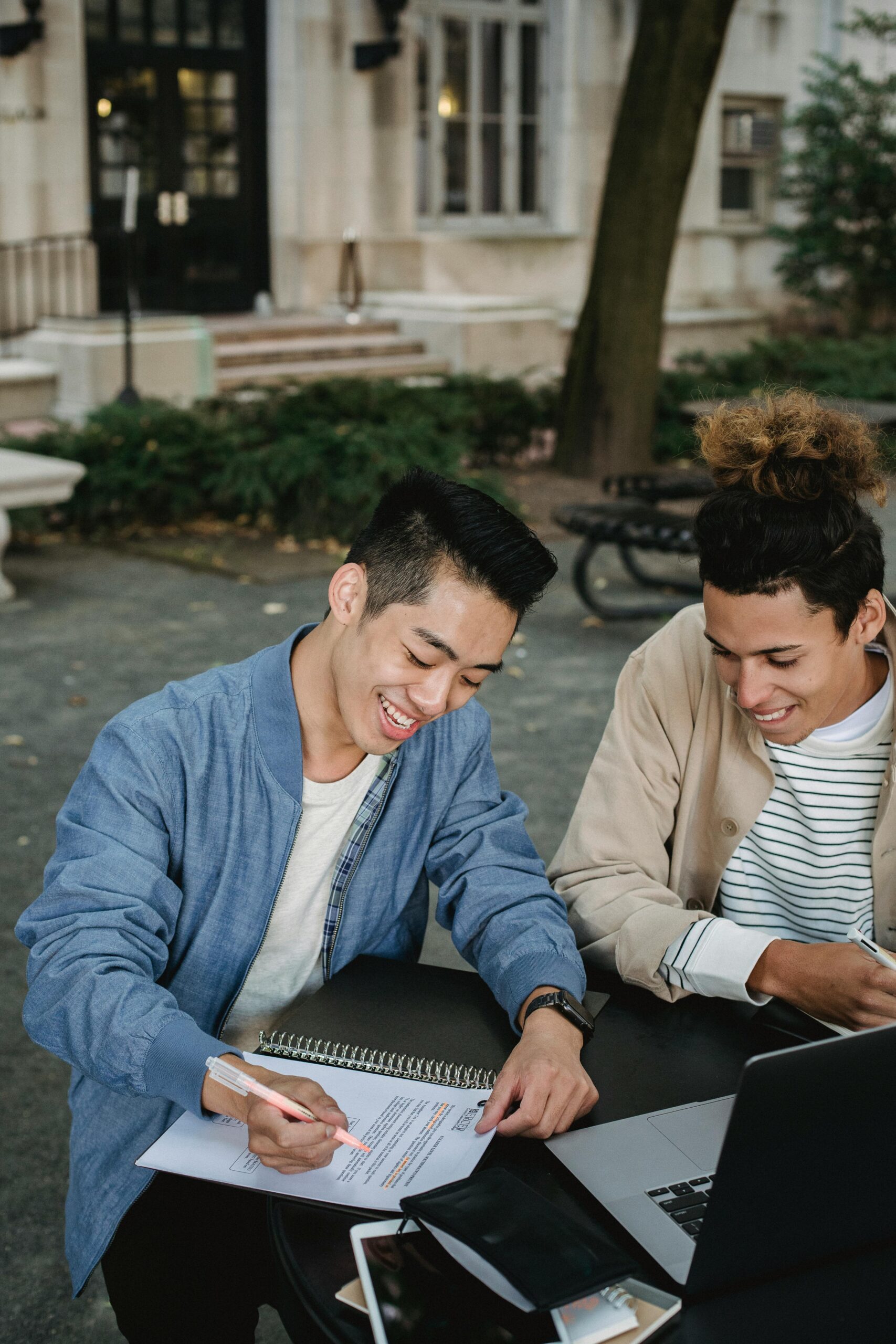 Two young men sit at an outdoor table, studying together with an open laptop, notebook, and papers in front of them.