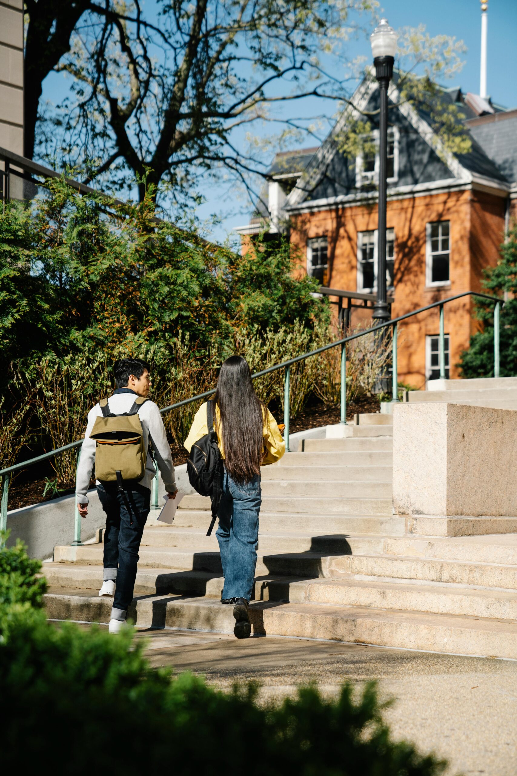 Two students with backpacks walk up outdoor steps on a campus, surrounded by greenery and buildings on a sunny day.