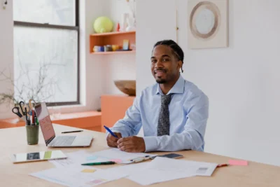 A man in a dress shirt and tie sits at a desk with paperwork, a laptop, and office supplies, smiling at the camera in a bright office space.