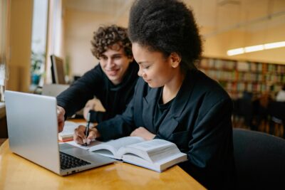 Two students study together at a table in a library, using a laptop and an open textbook. One points at the laptop screen while the other takes notes.