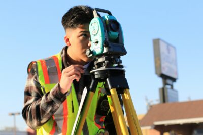 A person wearing a reflective safety vest uses a theodolite on a tripod to survey outdoors on a clear day.